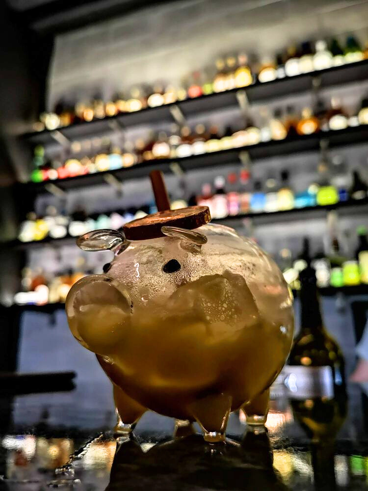 Cocktail in a clear glass container shaped like a piggy bank in front of the bar with blurred bottles of liquor in the background.