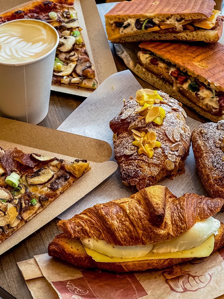 Coffee cup with latte art, slices of pizza, paninis, croissants, and croissant breakfast sandwich with poached eggs and cheese.